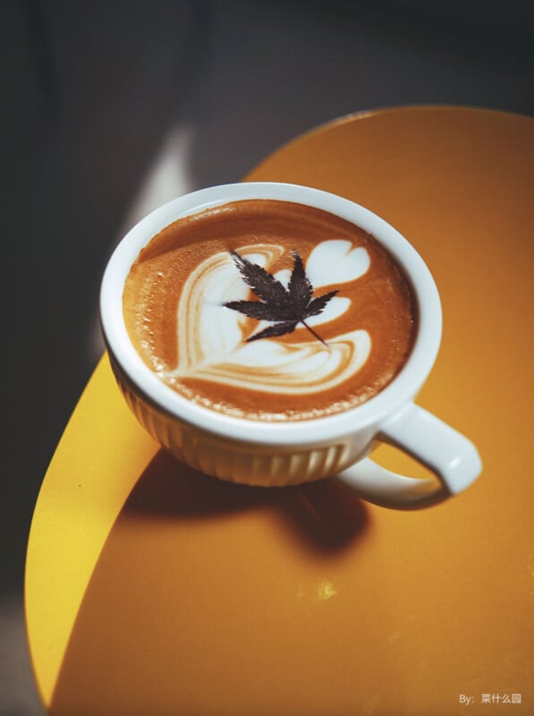 A cup of coffee with intricate latte art featuring a heart design and a small brown leaf on top. The cup is placed on a yellow table, casting a soft shadow.