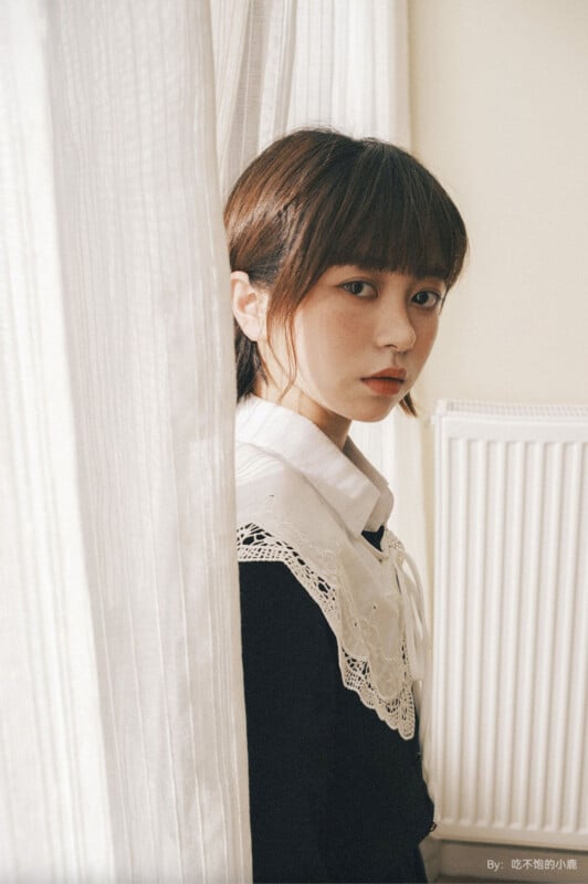 A young woman with short brown hair stands by white curtains, wearing a black outfit with a white lace collar. She gazes intently at the camera, with a radiator visible in the background.