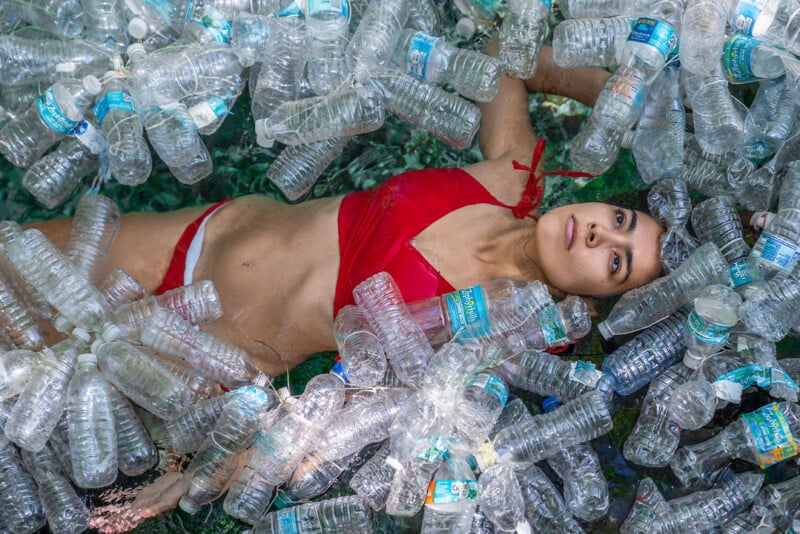 A person in a red bikini floats on their back in water surrounded by numerous empty plastic bottles. The bottles create a dense layer around them, highlighting environmental pollution. The water is visible through small gaps between the bottles.