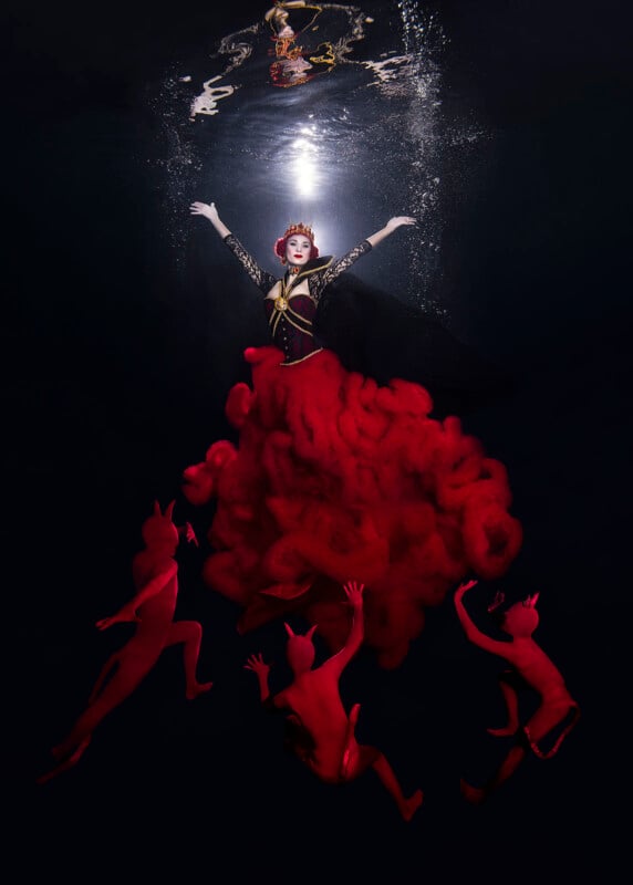 A person underwater wearing a dramatic red and black outfit with a glowing headpiece. Three small figures with red skin and horns swim around them. The person floats toward a light at the water's surface.