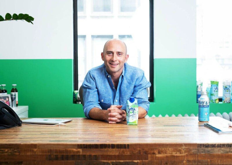 A person in a blue shirt smiles while leaning on a wooden desk. Various beverage bottles are displayed around them. The room has a green and white wall with a window in the background.