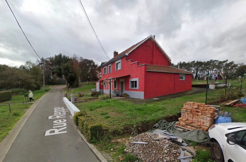 A red-brick house with a tiled roof sits on a grassy corner lot. A road sign reads "Rue Repose." A pile of bricks and a parked white car are in the foreground. Overcast skies loom overhead.