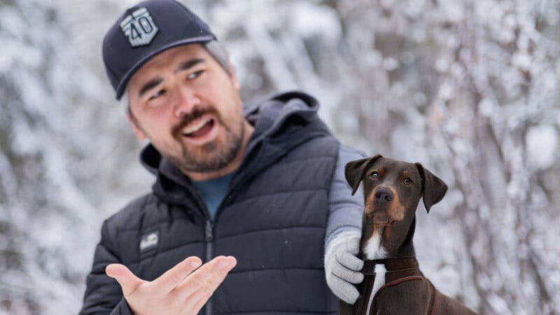 A person in a dark jacket and cap gestures animatedly while standing in a snowy landscape. A brown dog with a curious expression sits beside them. Snow-covered trees are blurred in the background.