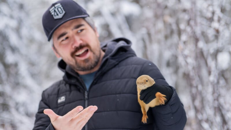 A man in a black jacket and cap gestures while holding a small animal in his gloved hand. Snowy trees are blurred in the background, suggesting a wintry outdoor setting.