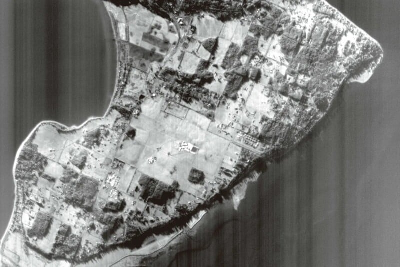 Aerial black and white image of a coastal area with patches of green fields, forests, and water bodies. The landscape is divided into irregularly shaped sections, showing a contrast between open fields and wooded areas along the shoreline.