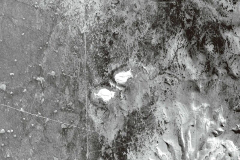 A grayscale aerial view showing a rugged, mountainous landscape with two bright, circular patches in the center. The terrain is uneven, with visible ridges, valleys, and variations in texture and shading.