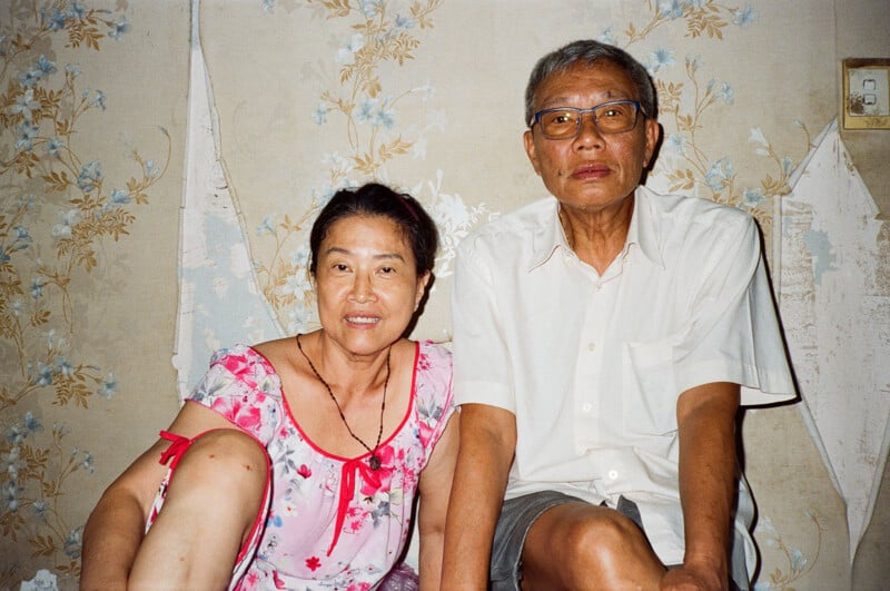 An older couple sits together in front of a wallpapered wall with floral patterns. The woman wears a floral dress, while the man is in a light-colored shirt and glasses. The wall shows signs of peeling.