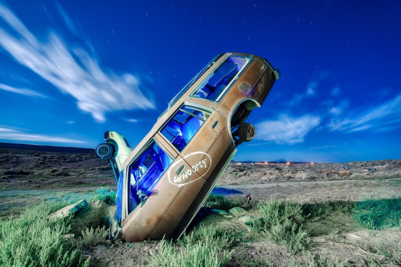 A car is partially buried nose-down in a grassy landscape under a starry night sky with streaking clouds. The car is positioned vertically, with its headlights facing the sky. Its interior glows with a blue light, and "Odyssey" is painted on its side.