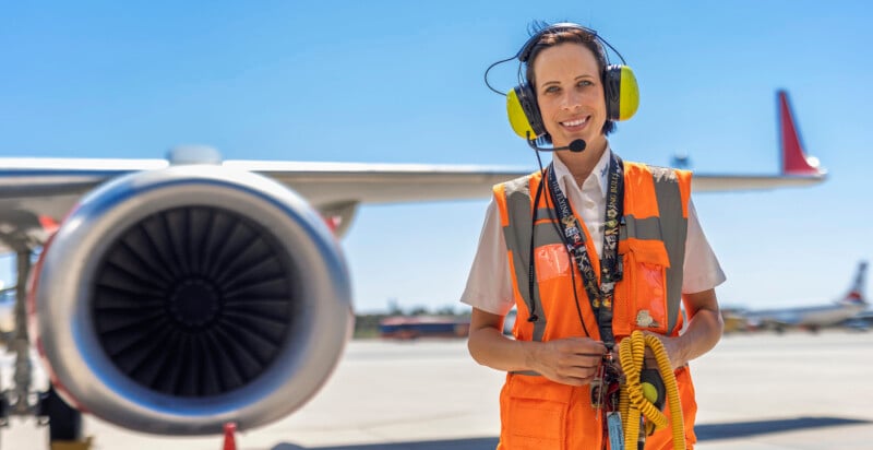 A woman in an orange safety vest and yellow ear protection stands on an airport tarmac beside a plane engine. She is holding coiled cables and wearing a headset. The sky is clear and blue.