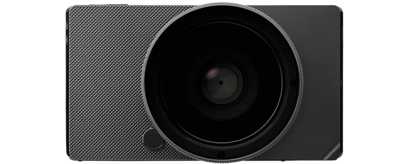 Close-up of a black digital camera with a textured front and a large circular lens at the center. The lens dominates the design, and the camera has a minimalist appearance.