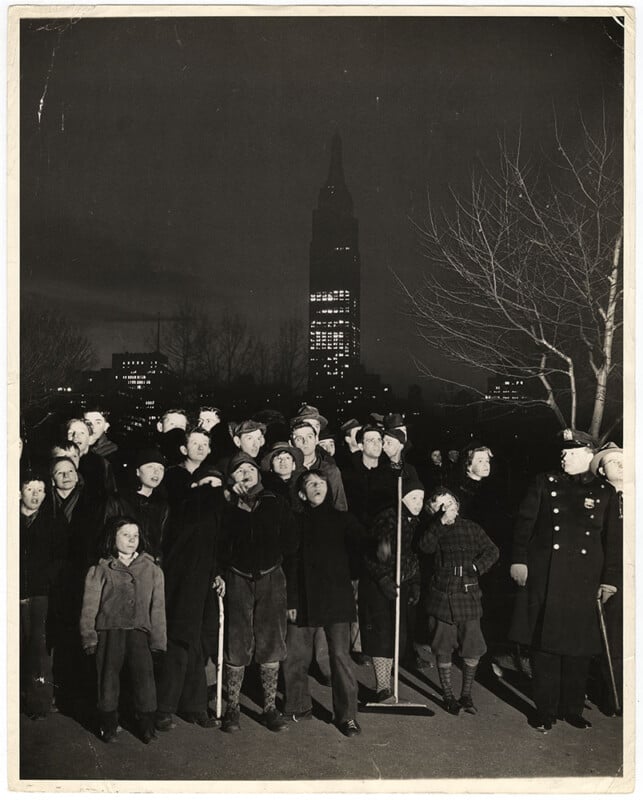 A group of people, including children, stand outdoors at night, looking upward with expressions of wonder. Two uniformed officers stand on the right. The silhouette of a tall building with lit windows is visible in the background.