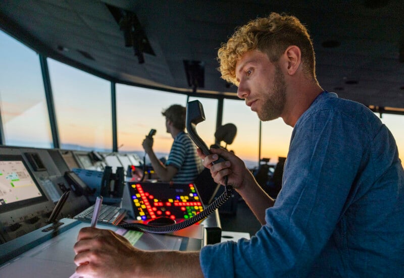 A man in a control tower holds a microphone and writes on a notepad, surrounded by control panels with colorful buttons. Another person is working in the background. The window shows a sunset sky.