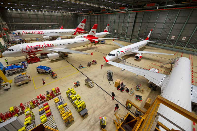 A spacious aircraft hangar with several red and white airplanes, including Austrian Airlines planes. Workers and equipment are visible on the floor. Bright overhead lights illuminate the scene.