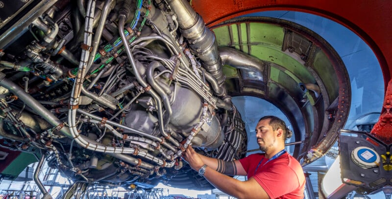 A person in a red shirt is working on a complex array of wires and pipes, possibly part of an aircraft engine. The person is focused, and the background shows parts of machinery and equipment.