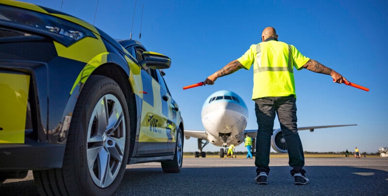 A ground crew member in a yellow vest uses red batons to guide an approaching airplane on a sunny day. An airport service vehicle is parked nearby on the runway.