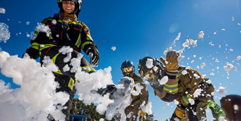 Firefighters are playfully interacting with foam during a training session. They are wearing full protective gear, including helmets and reflective stripes. The sky is clear and blue, and the scene is filled with motion and energy.