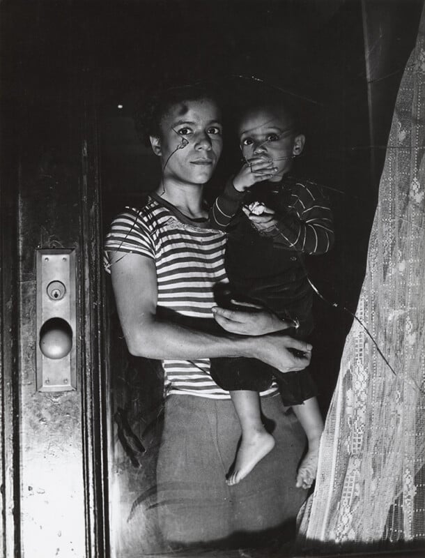 A person in a striped shirt holds a small child in one arm, standing by a door partially covered with lace curtains. The child is barefoot and resting a hand near their mouth. The lighting casts strong shadows on their faces.