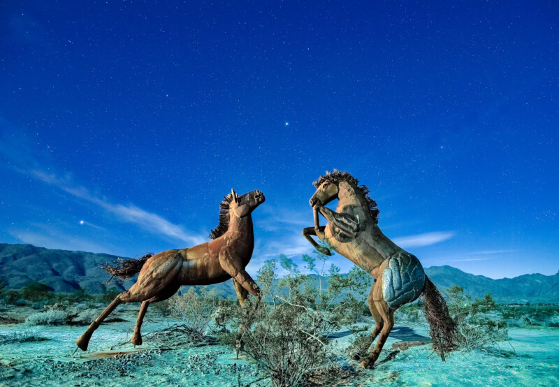Two large horse sculptures made of metal stand on a desert landscape under a starry night sky. One horse is rearing up, while the other stands on all fours. Mountains are visible in the background.