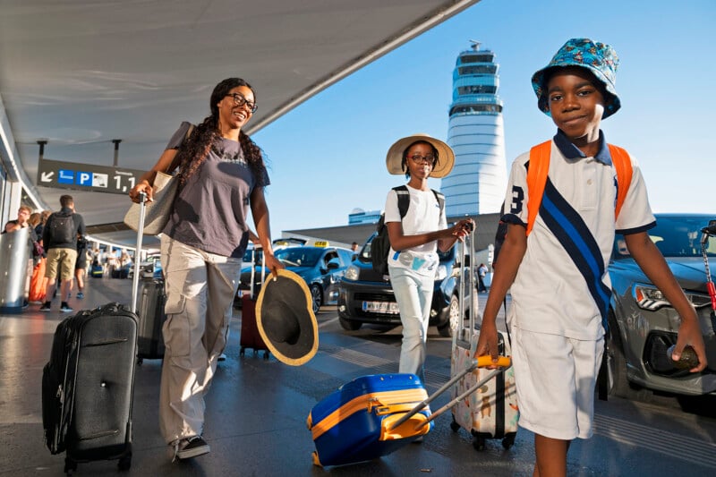 A smiling woman and two children walk through an airport with suitcases. They are wearing casual travel clothes, and one child is pulling a blue and orange suitcase. The background shows airport signage and parked cars.