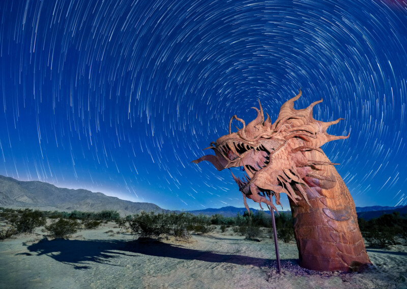 A metal dragon sculpture is silhouetted against a night sky filled with swirling star trails. The sculpture stands in a desert landscape, with mountains visible in the background. The long exposure creates a sense of motion in the stars.