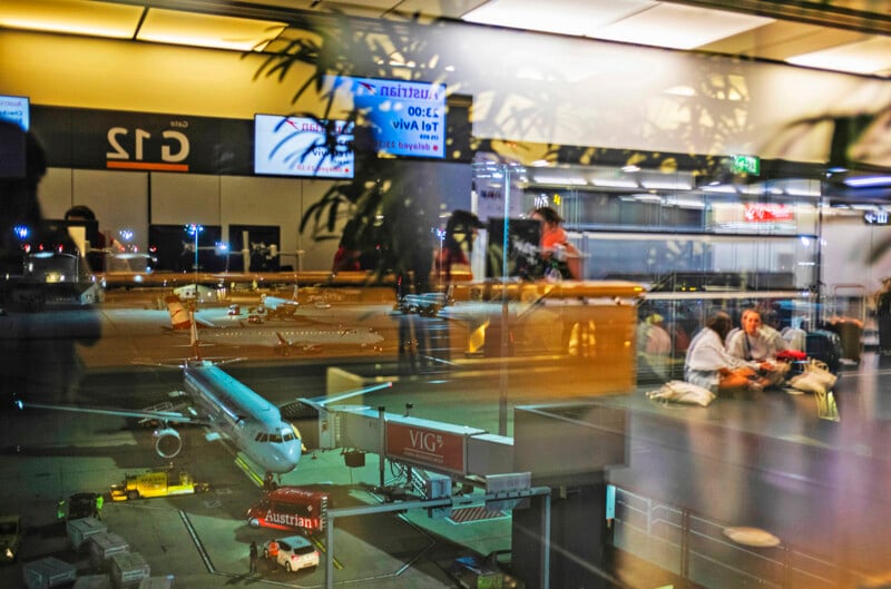 View of an airport gate from a terminal window. A plane is parked outside, while inside, passengers are seated and waiting. Reflections of lights and plants create a layered effect between inside and outside scenes.