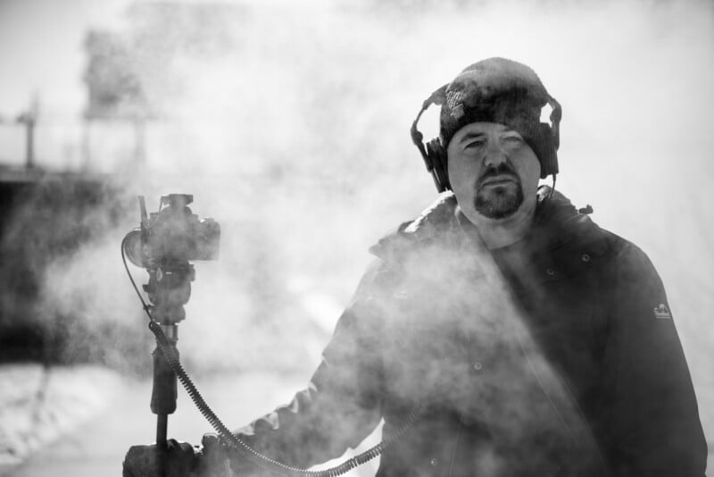 A man in a winter jacket and beanie stands outdoors, surrounded by fog or mist. He wears headphones and holds a camera on a pole with a cable attached. The scene is in black and white, giving a moody effect.