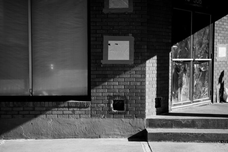 Black and white photo of a brick building corner with a covered window and glass doors. Sunlight casts a diagonal shadow on the structure. A small step leads to the entrance. The overall scene appears empty and quiet.