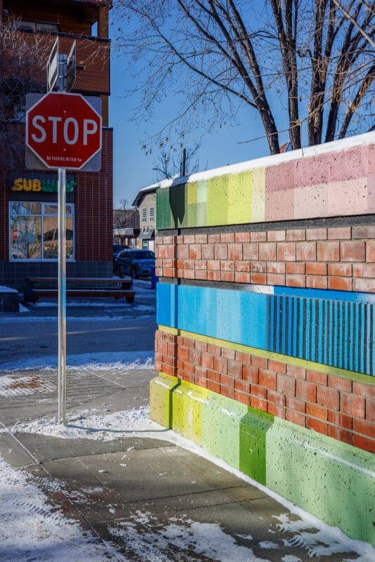 Stop sign on a snowy sidewalk near a colorful brick wall with green, blue, and pink horizontal stripes. Trees without leaves and a building with a "Subway" sign are visible in the background under a clear blue sky.