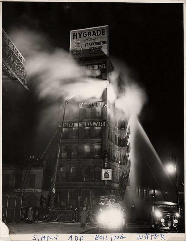 Firefighters battle a blaze in a multi-story building at night. Smoke and water spray fill the air. A vintage truck is in the foreground. A billboard at the top advertises Hygrade Frankfurters. Handwritten text reads "SIMPLY ADD BOILING WATER.