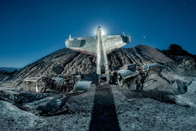 A dilapidated airplane rests abandoned on a rugged, rocky landscape under a starry night sky. The moonlight casts a silhouette of the tail section on the ground, and barren hills form the background.