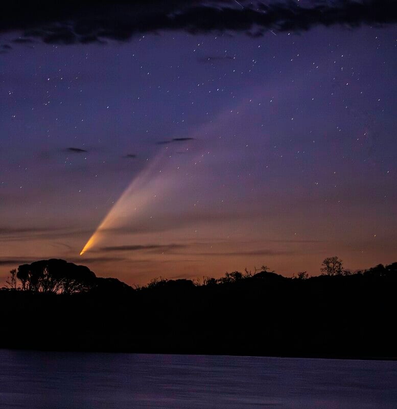A comet with a bright tail streaks across a starry night sky above a silhouetted landscape. The foreground features a body of water and dark outlines of trees and hills.