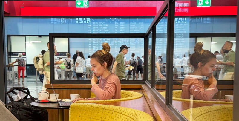 A woman sitting in a yellow chair, talking on the phone and using a laptop in a café. Reflected glass shows people walking in a busy indoor area. Red signage above lists news updates. Cups and a jacket are on the table.