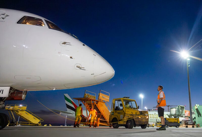 Airport ground crew works under floodlights beside a parked airplane at night. Workers in yellow uniforms handle equipment near the nose of the aircraft, while a worker in an orange vest stands nearby. Various airport vehicles are parked around.