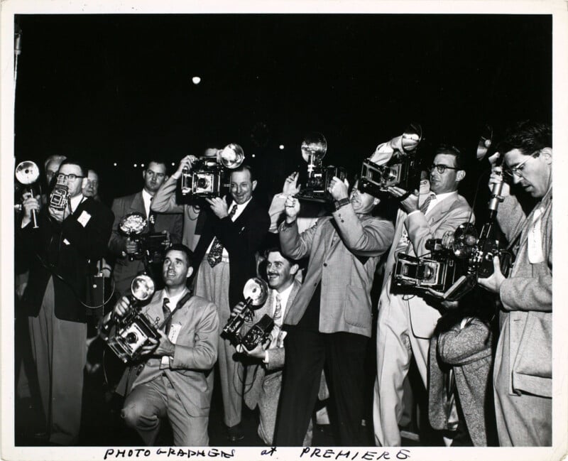 A group of photographers with vintage cameras are gathered at an event. They are dressed in suits and appear focused, capturing photos, creating an atmosphere of excitement and anticipation. The caption reads "Photographers at Premiere.