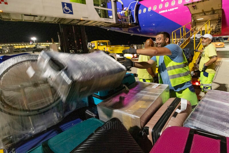 Airline workers in high-visibility vests load luggage, including wrapped suitcases and a bicycle, onto a conveyor belt at night. An aircraft with visible passenger seating is in the background.