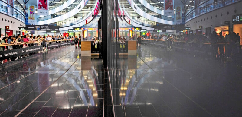 A busy airport terminal with rows of seated passengers and travelers lining up. The shiny tiled floor reflects the bustling scene and information signs above. Large windows arch overhead, allowing natural light to fill the spacious area.