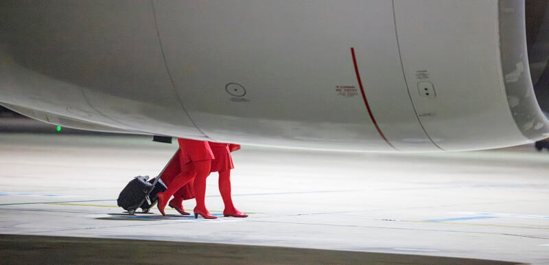 A person dressed in red, including a coat and high-heeled boots, is walking on an airport tarmac with a wheeled suitcase. The top of the person is obscured by an airplane's fuselage. The scene is dimly lit, suggesting evening or night.