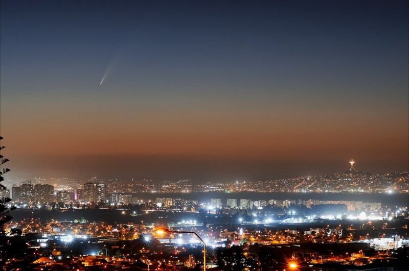 A cityscape at dusk with bright lights illuminating the buildings. In the sky, a comet with a glowing tail is visible. The horizon shows a gradient from orange to dark blue, and a notable illuminated statue stands on a distant hill.