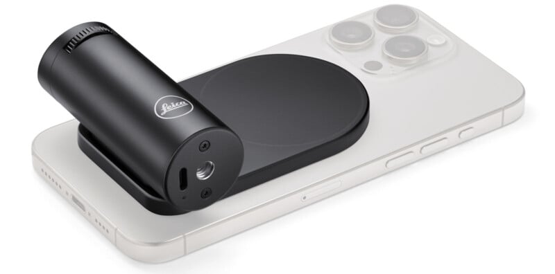 A black Leica attachment is positioned on a white smartphone with a triple camera system. The phone is lying flat, and the attachment is cylindrical and sleek, placed near the phone's camera area.