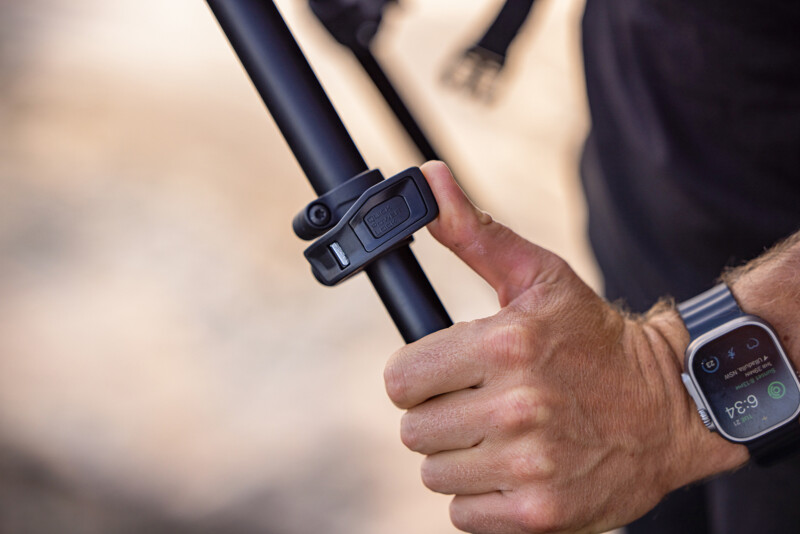 A person wearing a smartwatch adjusts a black adjustable pole, possibly part of a tripod or similar equipment, using a locking mechanism. The background is blurred, emphasizing the focus on the hand and the device.