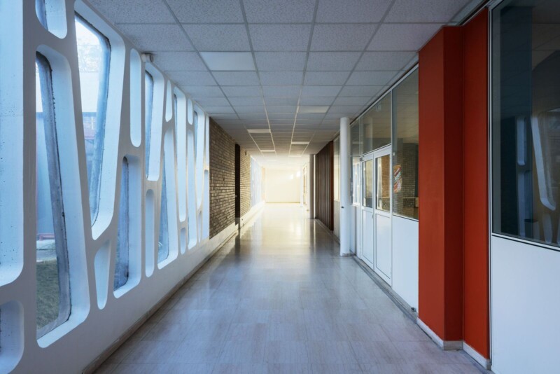 A modern hallway featuring geometric windows on the left, a brick wall on the right, and a red accent wall midway. The corridor is well-lit with a polished floor and a ceiling of white tiles, leading to a bright area at the end.