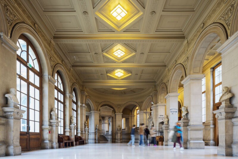 A grand hallway with ornate columns and sculptures along the walls. Large arched windows let in natural light, illuminating the high ceiling with intricate designs. People walk down the polished floor, creating a sense of movement in the elegant space.