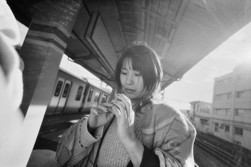 Black and white photo of a person with shoulder-length hair standing on a train platform. They are focused on using a smartphone. A train is visible in the background, along with station architecture. The scene captures a quiet, reflective moment.