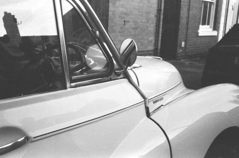 A black-and-white photo of a vintage car, showcasing its curved fender, side mirror, and branding badge. Brick buildings and another car are in the background, partially obscured.