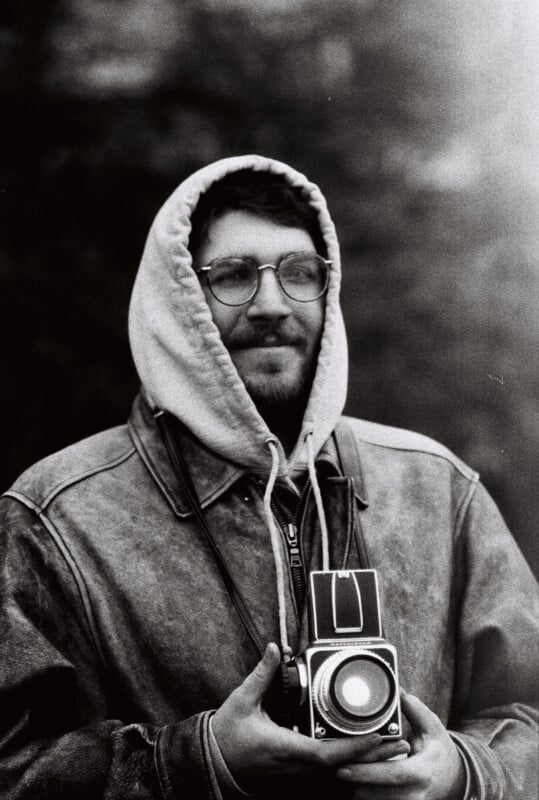 A black-and-white photo of a person wearing glasses, a hooded jacket, and holding a vintage camera. They are looking slightly to one side. The background is blurred, emphasizing the person and the camera in the foreground.