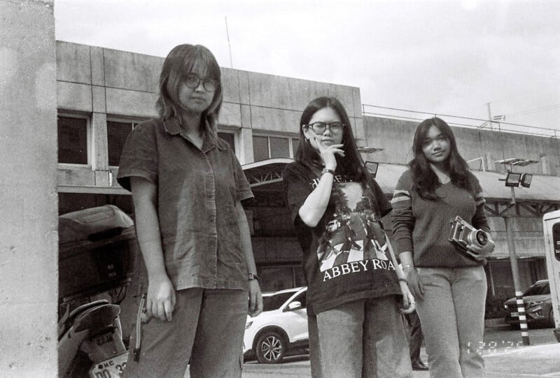 Three people stand outside a building. One wears glasses and a shirt, another wears glasses and an "Abbey Road" T-shirt while holding a cigarette, and the third holds a camera. Cars and part of a building are visible in the background.