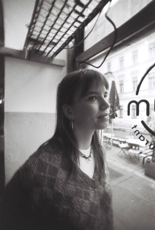 Black and white photo of a woman with long hair and bangs, wearing a sweater and earrings. She is looking to the right, standing near a window with the view of an outdoor area and buildings in the background.