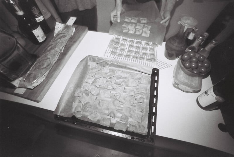 Black and white image of a kitchen scene. Unbaked ravioli arranged on a parchment-lined tray atop a counter. Nearby, a bottle of wine, fresh tomatoes, and various ingredients are visible. Two people are preparing food in the background.