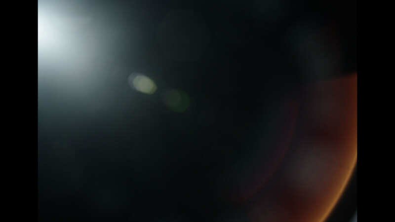 A dark, abstract image with a bright light source in the top left corner creating lens flares, and subtle hues of red and orange fading into black. The overall effect is mysterious and ethereal.