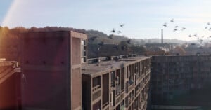 Aerial view of a large, rectangular, brick apartment building with birds flying overhead. In the background, a hilly landscape with scattered trees and a smokestack is visible under a clear sky.